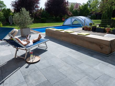 Donna che si rilassa su un lettino in una terrazza con lastre di calcare, sullo sfondo un giardino con piscina