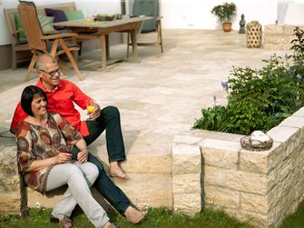Coppia seduta su una terrazza con lastre di pietra naturale in travertino di Medium Select e muro in pietra naturale, mobili da giardino in legno sullo sfondo