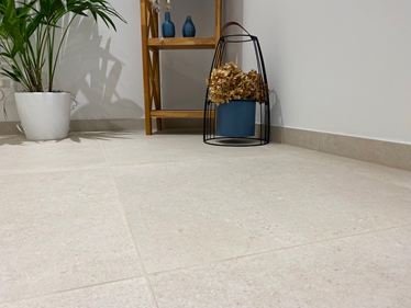 Primo piano di un pavimento con piastrelle in gres porcellanato Sevilla Cream dall'aspetto calcareo. Sullo sfondo una mensola in legno, una pianta e delle decorazioni.