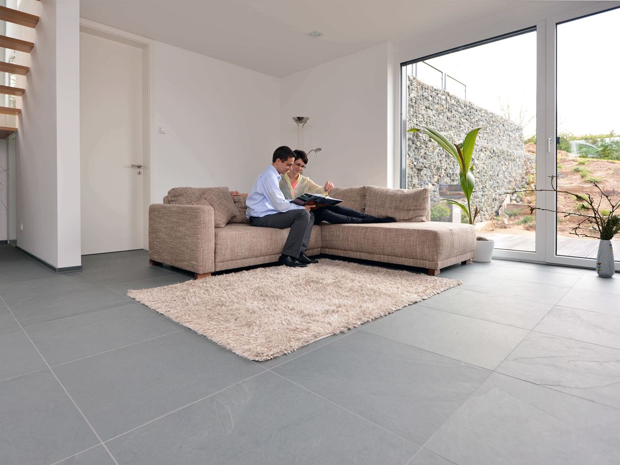 Grey Slate ha un aspetto spazioso e moderno. Piastrella in ardesia Grey Slate di grande formato nel soggiorno con divano chiaro e pareti bianche. Una coppia siede sul divano