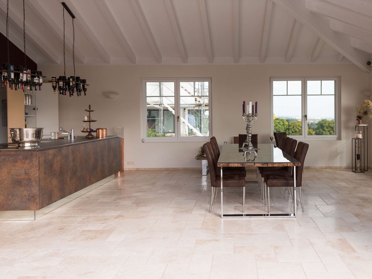 La cucina moderna incontra il piano mediterraneo. Piastrelle in travertino chiaro a legante romano in una cucina moderna con tavolo da pranzo e isola di cottura