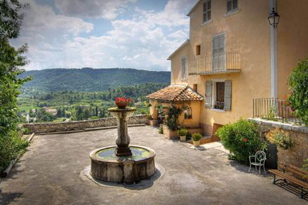 Una fontana in una piazza accanto a una casa di campagna, montagne sullo sfondo