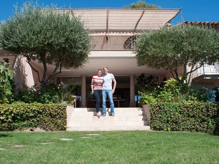 Coppia in piedi davanti a un hotel con pavimento in travertino, prato in primo piano e alberi a sinistra e a destra