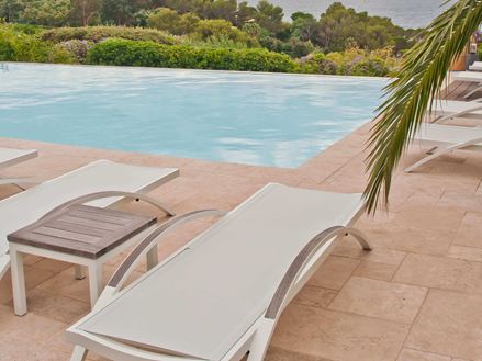 Sedie a sdraio bianche su un pavimento di travertino a bordo piscina, un ramo di palma sporge nell'immagine da destra