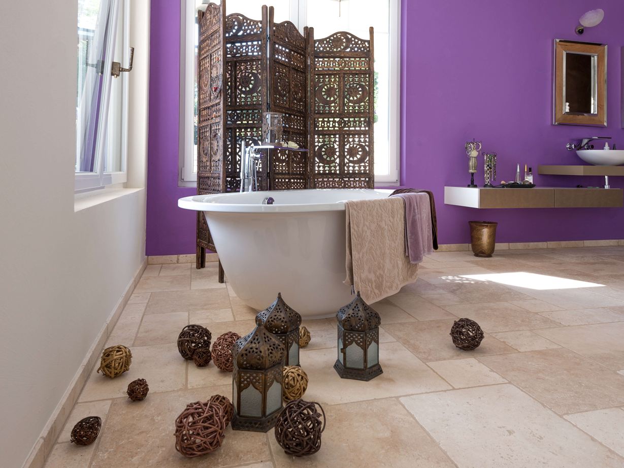 Piastrelle Light in travertino in un bagno con vasca freestanding e parete viola sullo sfondo