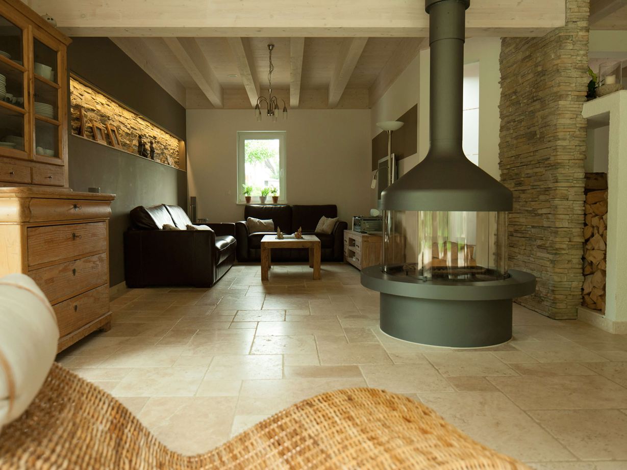 Soggiorno con camino autonomo e zona salotto su piastrelle di travertino Light, poltrona in vimini in primo piano