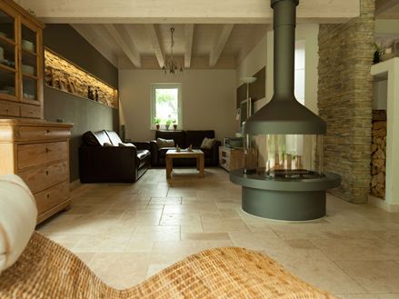 Soggiorno con camino autonomo e zona salotto su piastrelle di travertino Light, poltrona in vimini in primo piano