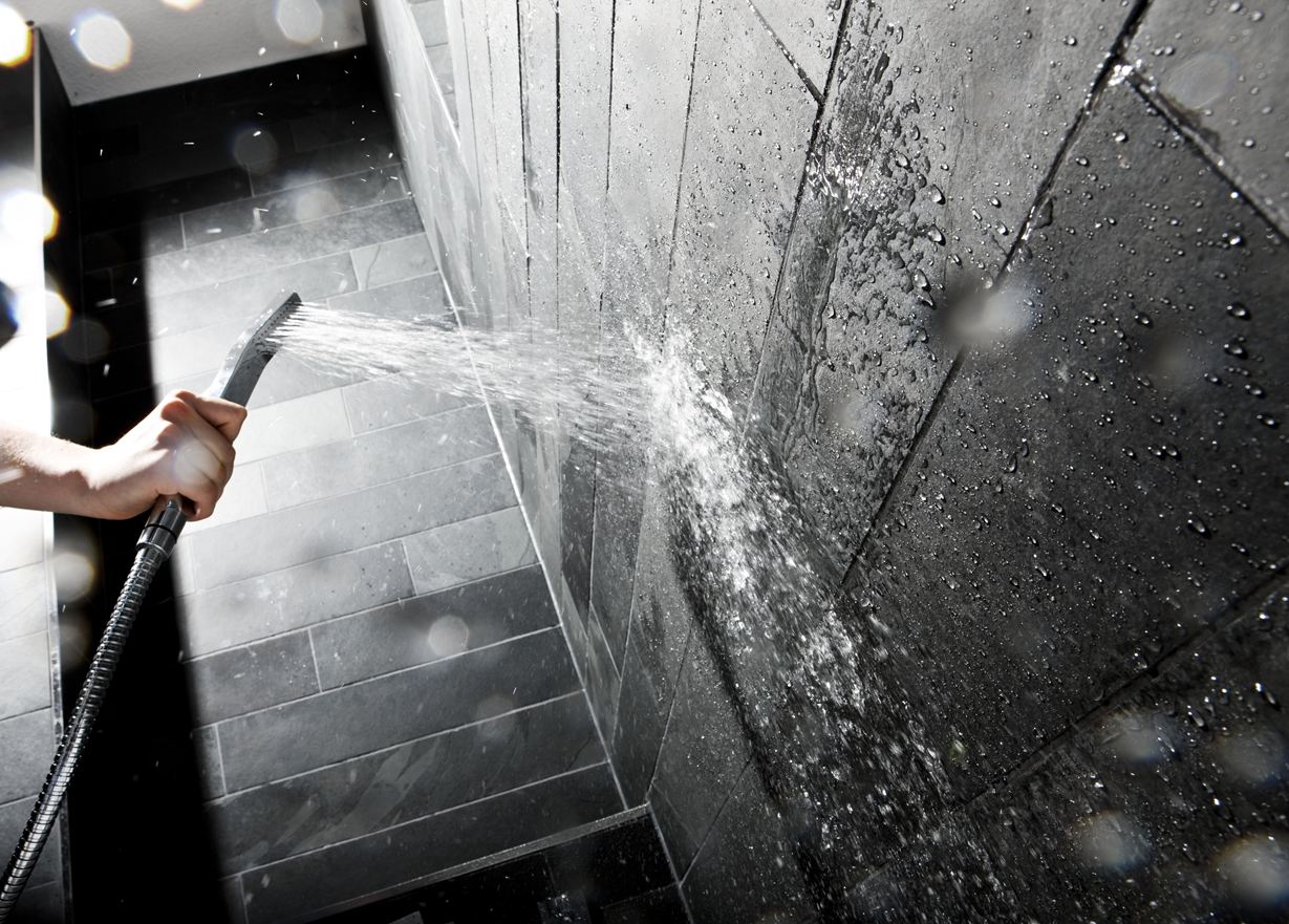 Mano che tiene il soffione di una doccia contro una parete con piastrelle di pietra scura
