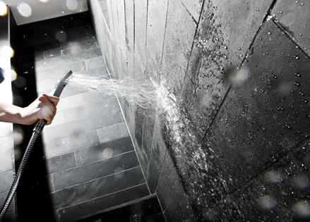 Mano che tiene il soffione di una doccia contro una parete con piastrelle di pietra scura