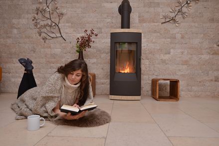 Donna sdraiata su piastrelle in pietra calcarea davanti a una parete con rivestimenti in pietra naturale e camino, mentre legge un libro